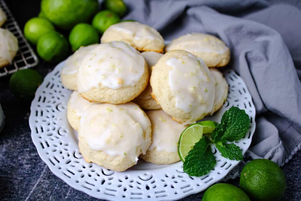 Plate of soft Key Lime Ricotta Cookies topped with sweet lime glaze, garnished with fresh limes and mint on a white serving platter.