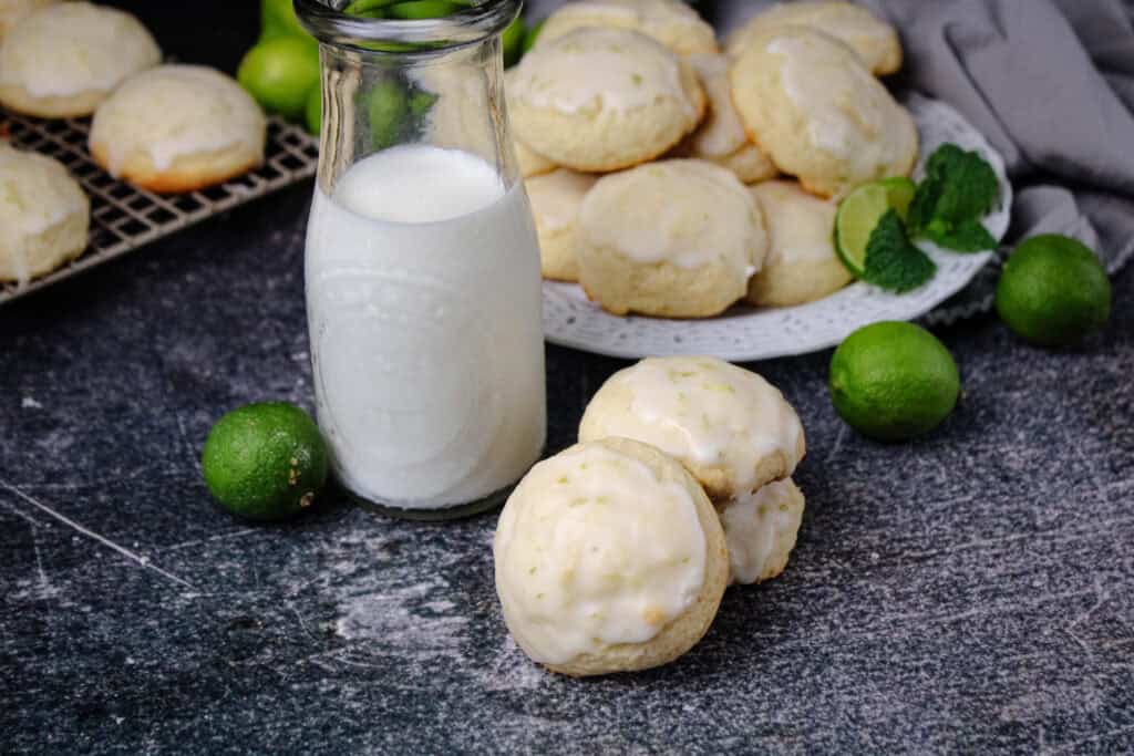 Stack of glazed Key Lime Ricotta Cookies served with a glass bottle of milk and fresh limes on a dark background.