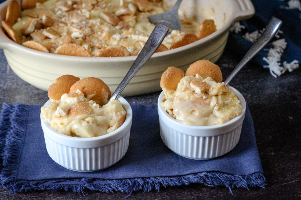 Two small ramekins filled with creamy homemade banana pudding topped with Nilla wafers, with a baking dish of banana pudding in the background.