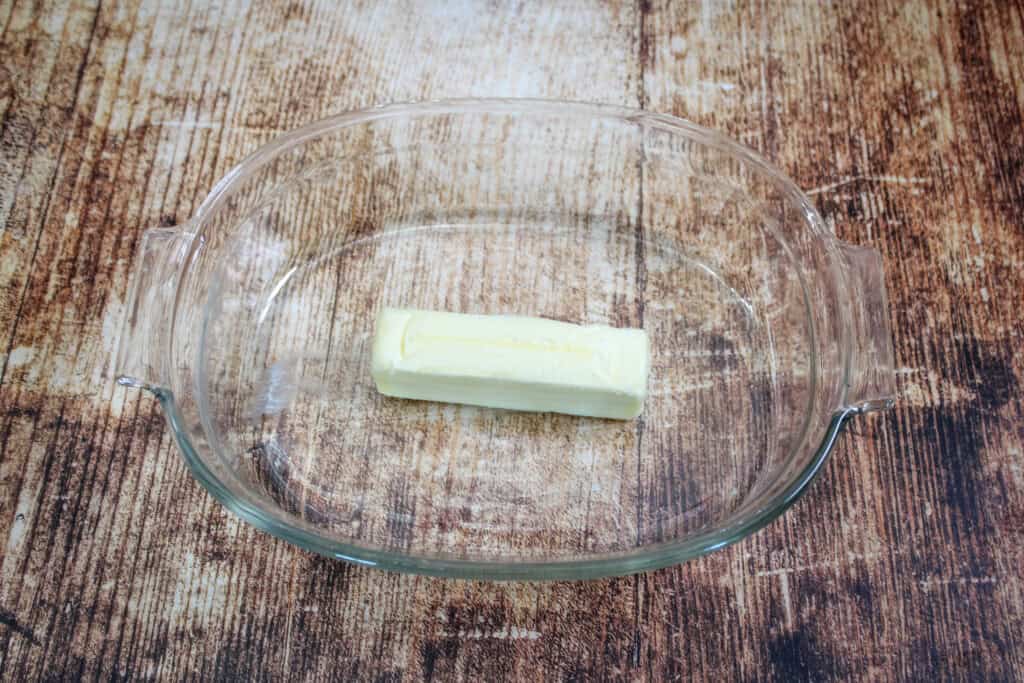 One stick of butter placed in a clear baking dish, ready to melt in the oven as the first step of making peach cobbler.

