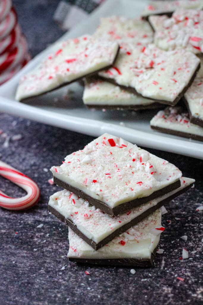 Three pieces of peppermint bark stacked next to a candy cane and a plate of them 