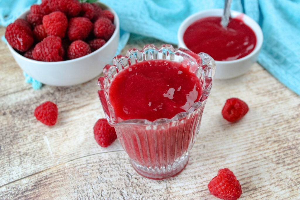 Raspberry sauce in a cup surrounded by fresh raspberries 