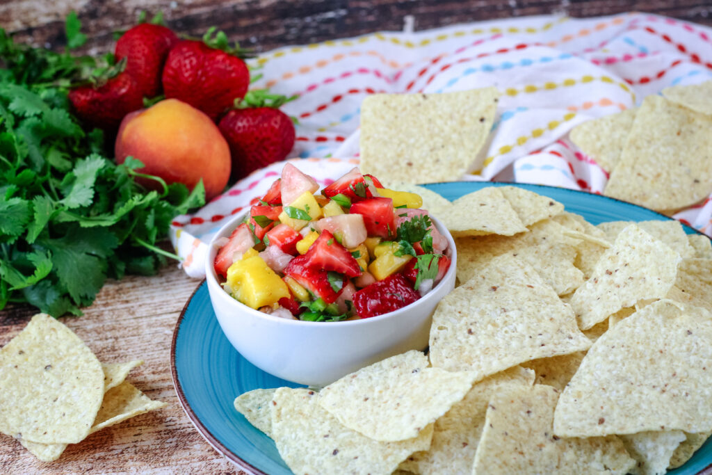 Fruit salsa in a white bowl placed on a turquoise plate with tortilla chips around it  with fruit and cilantro in the background