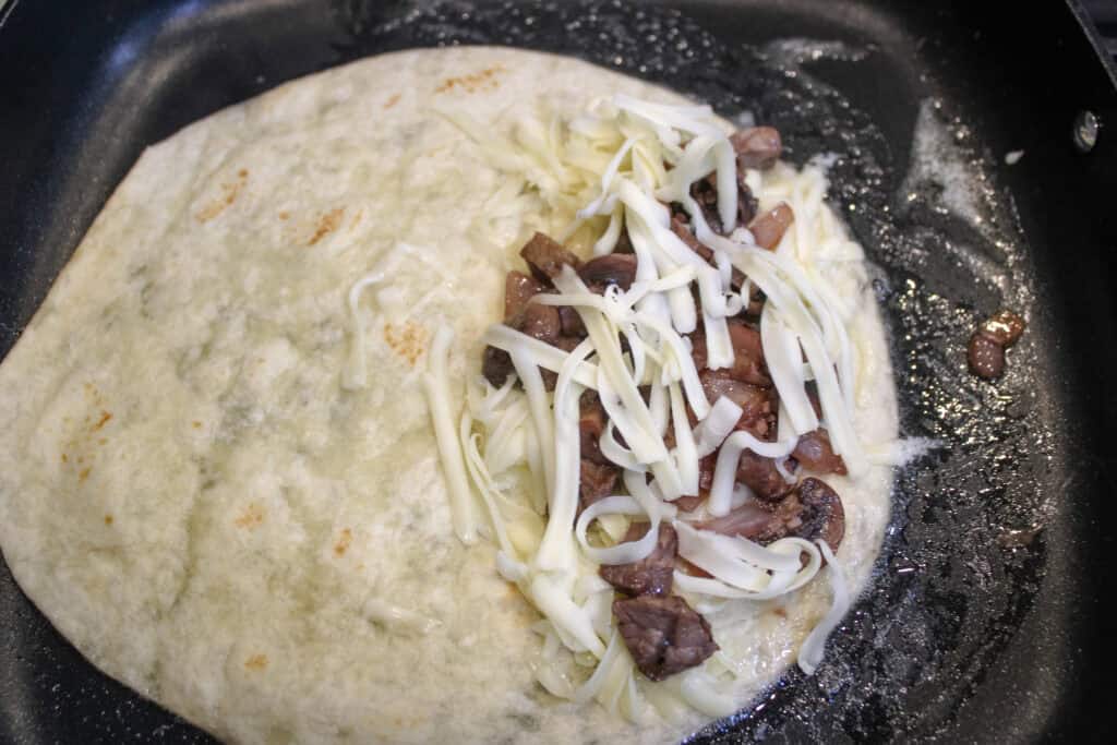 A tortilla in a skillet with cheese , steak , mushrooms , onions and garlic on one side