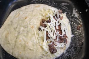 A tortilla in a skillet with cheese , steak , mushrooms , onions and garlic on one side