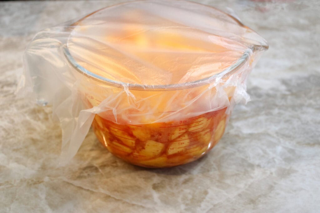 Peaches diced up in a measuring cup with sugar making their own syrup covered with plastic wrap , making peach jam