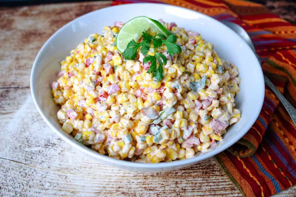 Close-up of a bowl of creamy corn salad garnished with a lime wedge and fresh cilantro, surrounded by smaller ramekins of the salad on a rustic table with a colorful striped napkin and vintage spoon.
