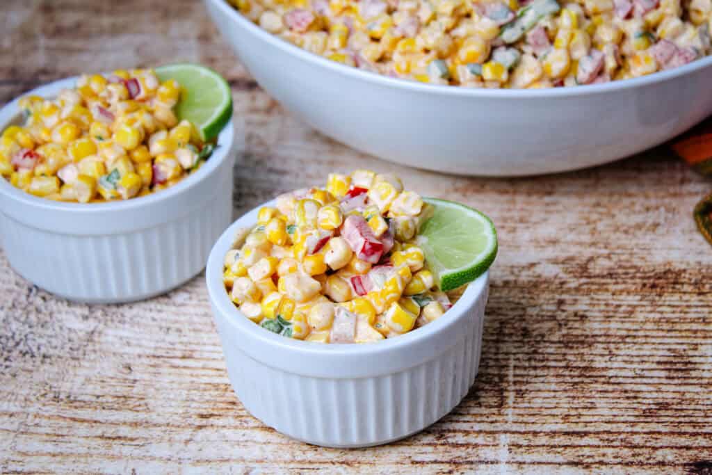 Close-up of creamy corn salad served in white ramekins, garnished with lime wedges. A large bowl of the same salad is visible in the background on a rustic wooden surface.