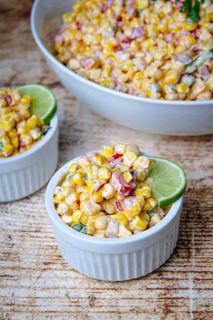 Close-up of creamy corn salad served in white ramekins, garnished with lime wedges. A large bowl of the same salad is visible in the background on a rustic wooden surface.