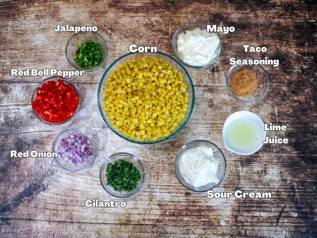 Flat lay of labeled ingredients for creamy corn salad on a rustic wooden surface, including corn, red bell pepper, red onion, jalapeño, cilantro, mayo, sour cream, taco seasoning, and lime juice in clear bowls.