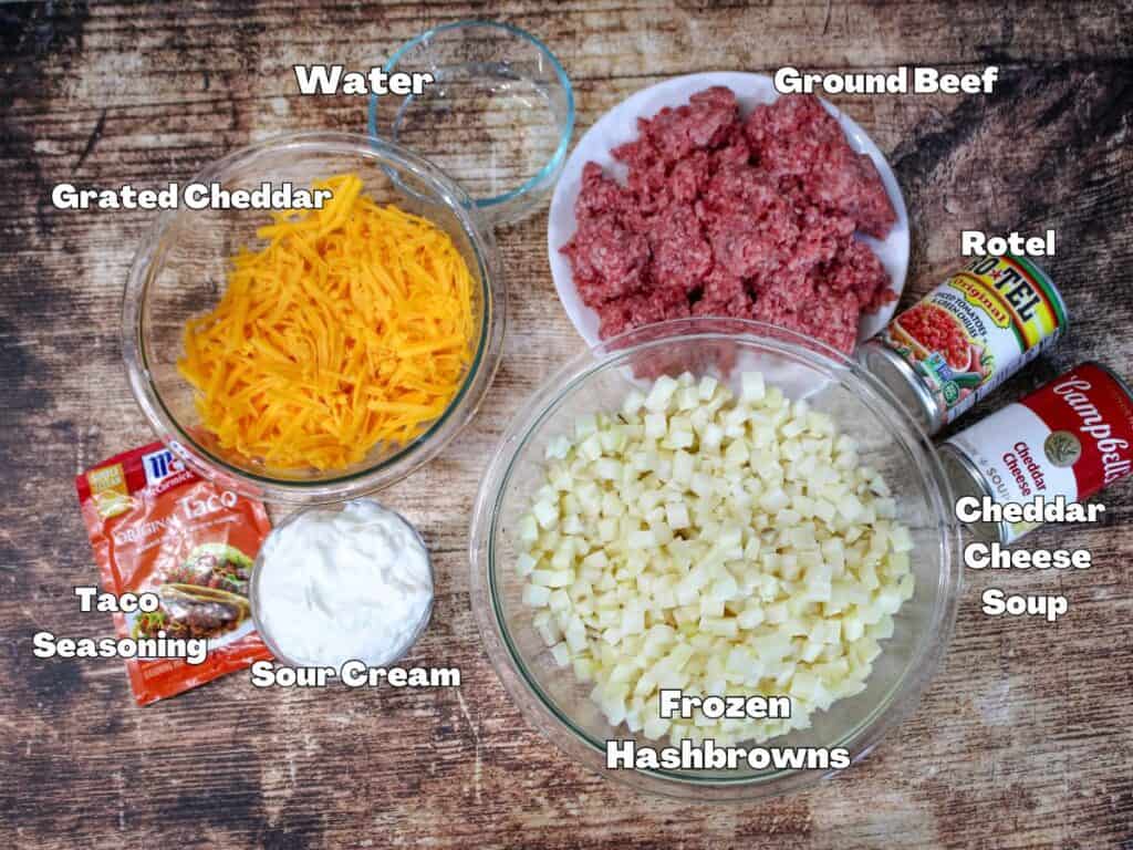 Ingredients for Taco Potato Casserole laid out on a wooden surface, including ground beef, Rotel, cheddar cheese soup, frozen hashbrowns, grated cheddar, taco seasoning, sour cream, and water.
