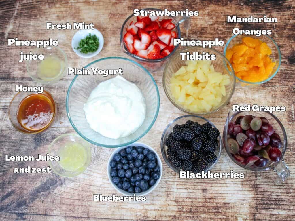 Flat lay of labeled ingredients for creamy fruit salad, including strawberries, pineapple tidbits, mandarin oranges, red grapes, blackberries, blueberries, plain yogurt, honey, lemon juice and zest, pineapple juice, and fresh mint on a rustic wooden background.