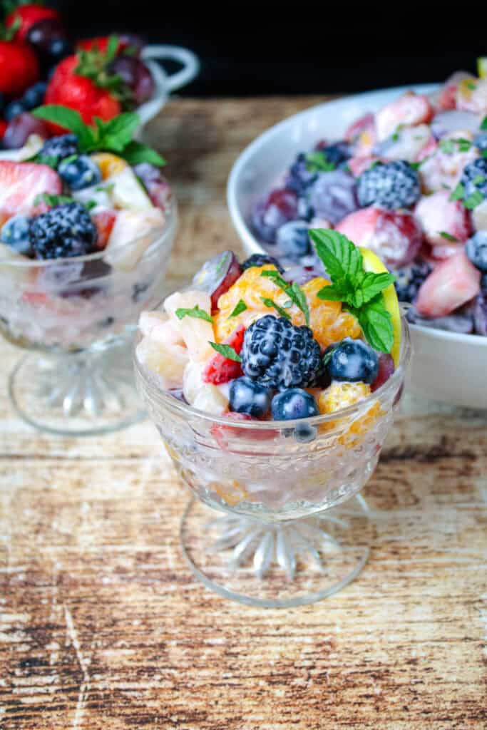 Individual glass dessert cups filled with creamy fruit salad made with blackberries, blueberries, mandarin oranges, strawberries, and fresh mint, with a large serving bowl in the background.