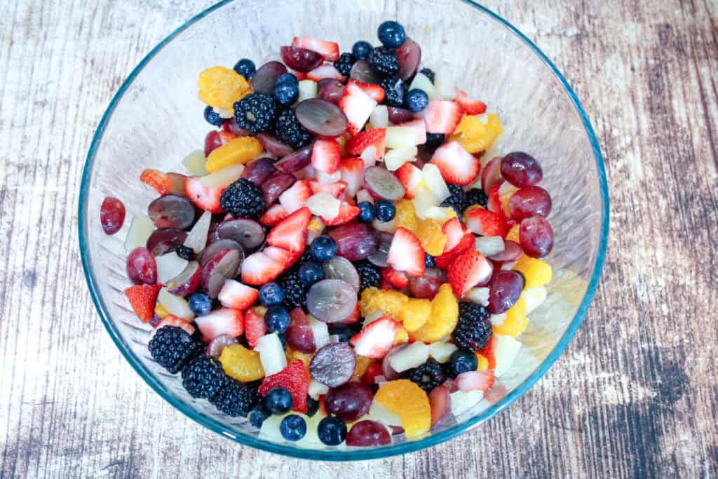 Glass mixing bowl filled with chopped fresh fruit including strawberries, blueberries, blackberries, mandarin oranges, grapes, and pineapple for creamy fruit salad.