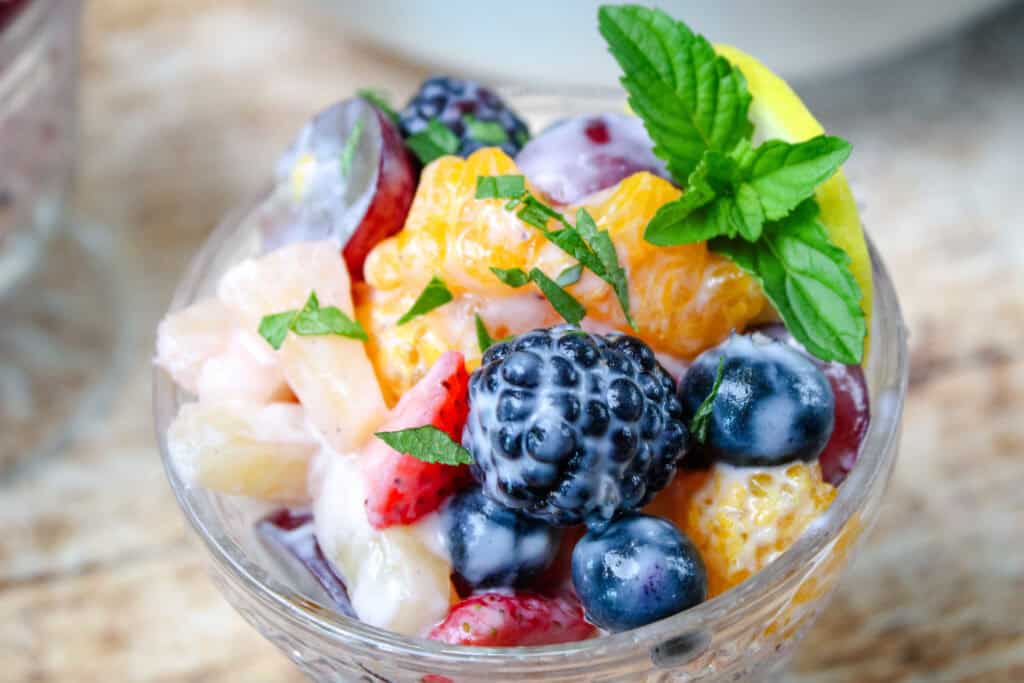 Close-up of creamy fruit salad served in a glass dish, topped with freshly chopped mint, lemon slice, and whole berries including blackberries, blueberries, and mandarin oranges.