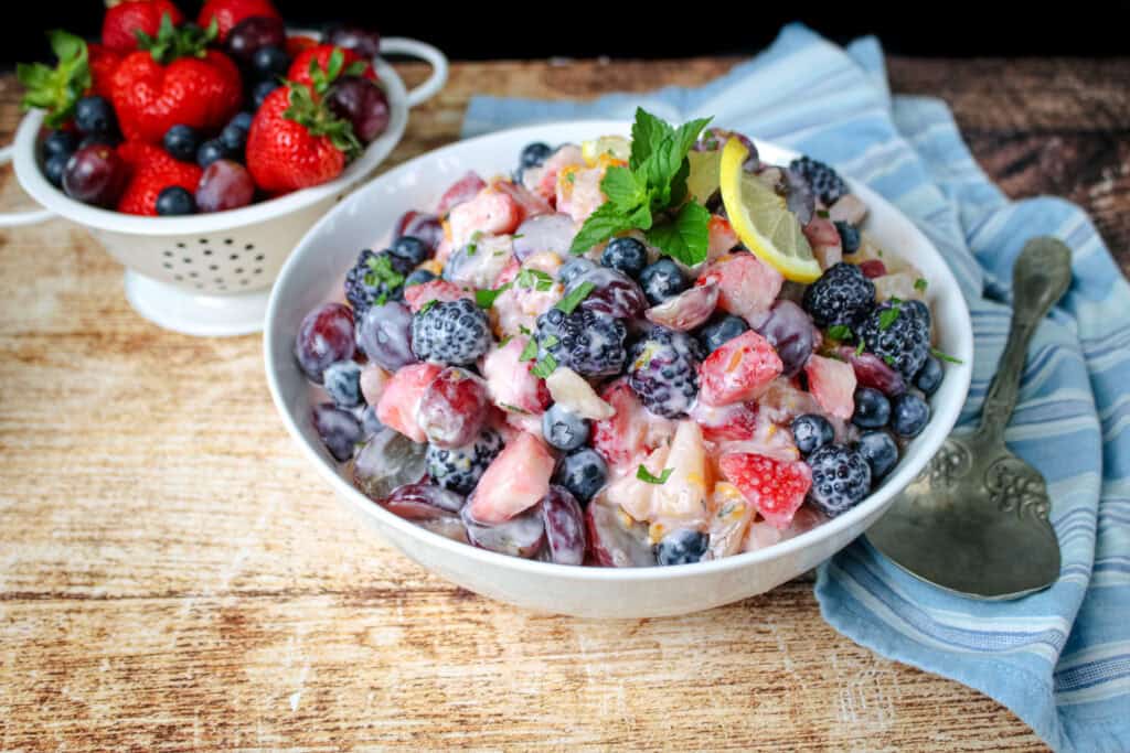 Large white bowl of creamy fruit salad garnished with fresh mint and lemon slices, surrounded by berries and served with a vintage spoon on a striped cloth napkin.