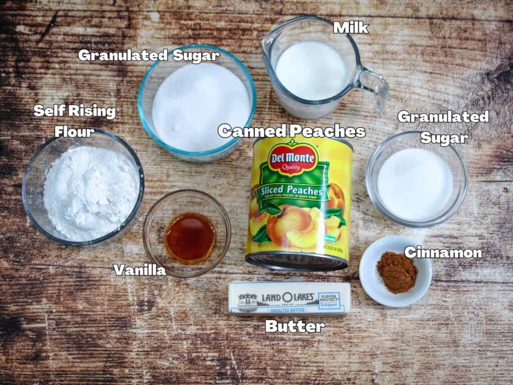 Overhead view of cobbler ingredients including canned peaches, self-rising flour, sugar, milk, butter, vanilla, and cinnamon labeled for recipe prep.