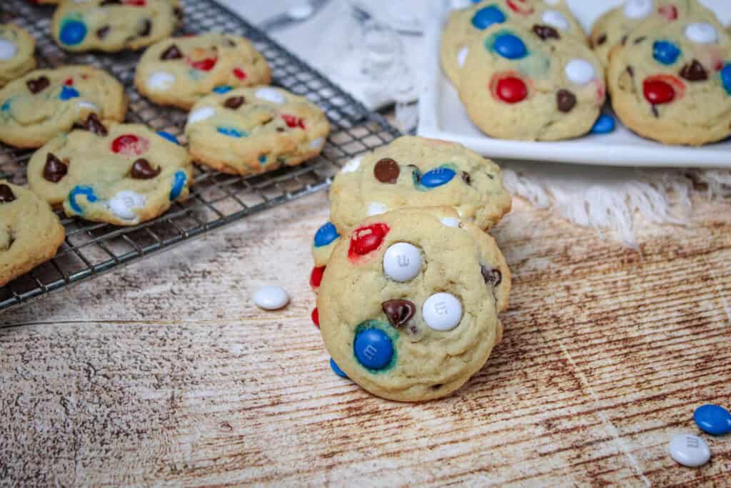 Close-up of baked Patriotic M&M Cookies resting on a cooling rack and rustic wooden surface, with M&M’s and chocolate chips clearly visible.