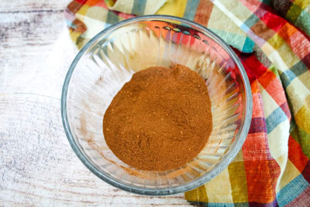 Clear glass bowl filled with homemade pumpkin pie spice blend on a rustic wooden surface, next to a plaid cloth in warm autumn colors.