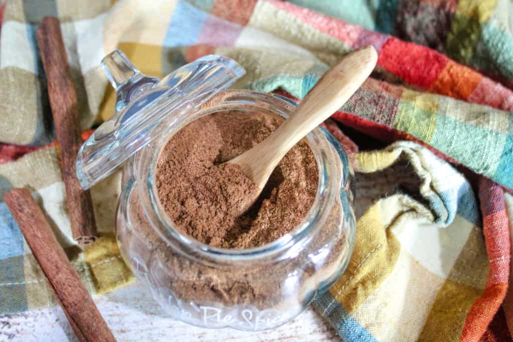 Open glass jar filled with homemade pumpkin pie spice, with a small wooden spoon resting inside, surrounded by cinnamon sticks and a colorful plaid cloth.