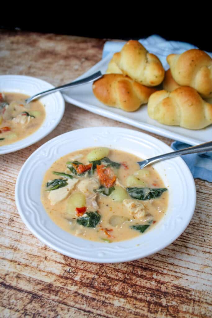 Two white bowls of Marry Me Chicken Soup with gnocchi, spinach, and sun dried tomatoes, served with golden garlic knots on a white platter in the background.