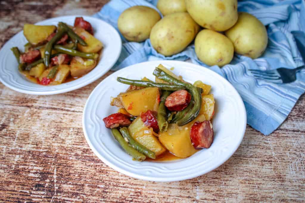 Two bowls of swamp potatoes featuring sausage, green beans, and golden potatoes with whole potatoes and a striped cloth in the background.