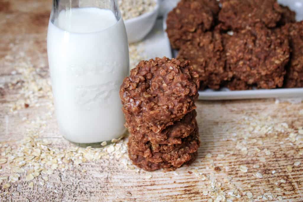 Close-up of chewy chocolate oatmeal cookies stacked next to a milk bottle, with a tray of cookies and oats in the background.