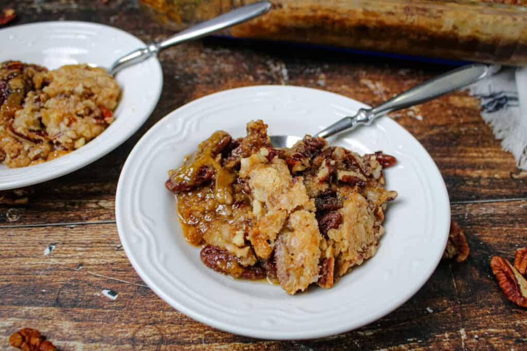 Two white dessert plates filled with warm pecan pie dump cake showing gooey caramel filling and crunchy pecans, with a baking dish in the background on a wooden table.