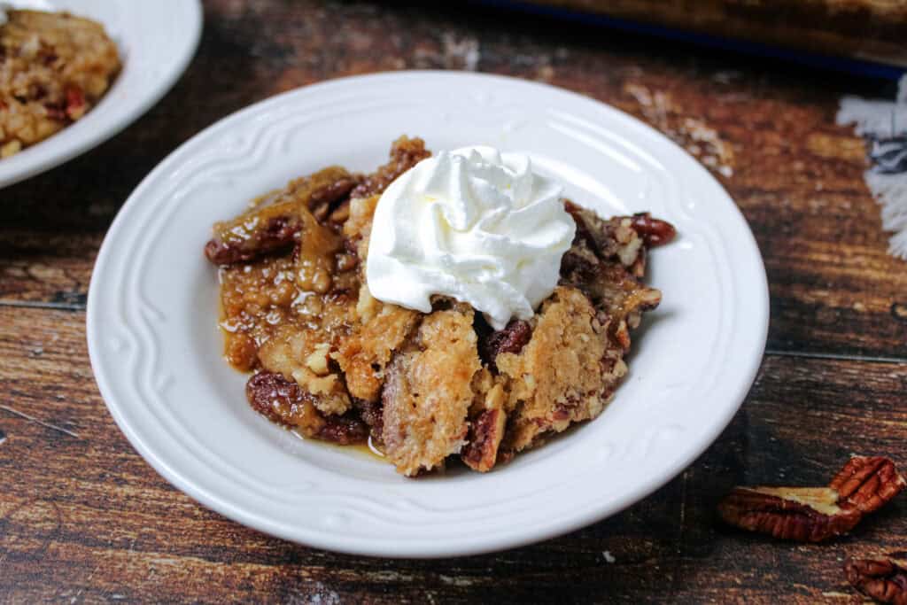 A serving of pecan pie dump cake topped with a swirl of whipped cream on a white plate, showing the rich, gooey layers of pecans and brown sugar filling underneath.