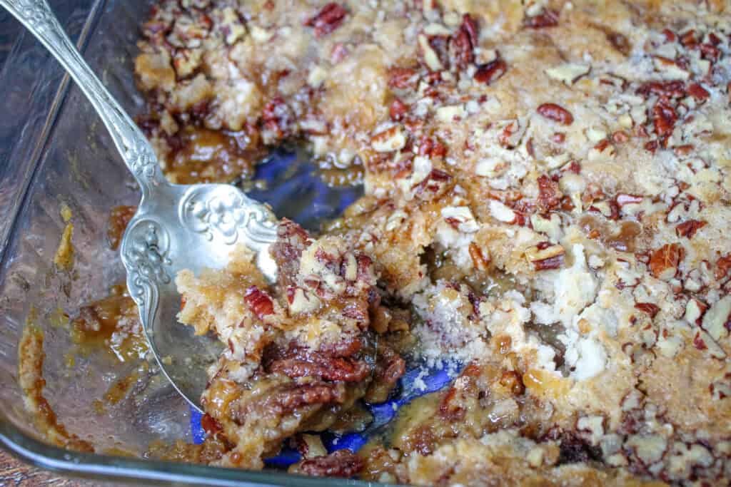 Close up of a serving spoon scooping pecan pie dump cake from the baking dish, revealing the sticky pecan filling and soft, golden cake like topping.