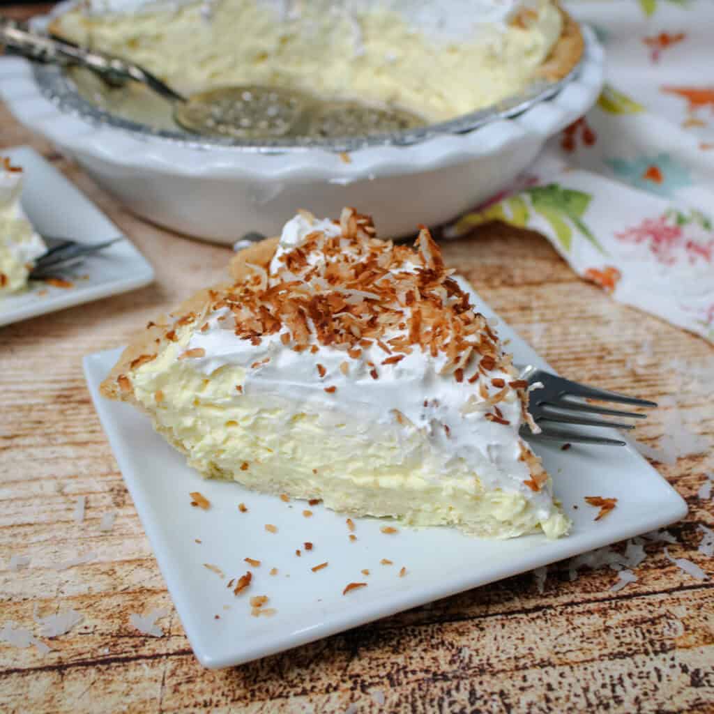 Slice of easy coconut cream pie topped with whipped topping and toasted coconut flakes on a white plate with fork, showing creamy coconut filling and flaky crust.